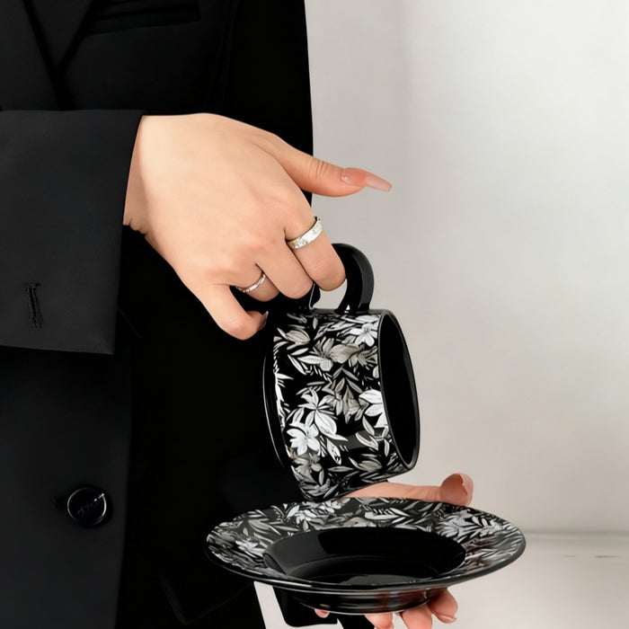 Person holding a black mug with floral pattern and matching saucer.